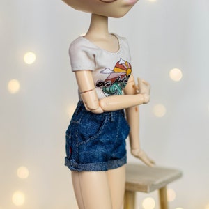 May include: A doll wearing a white t-shirt with a colorful graphic of a sun, mountains, and waves. The doll is also wearing blue denim shorts. The doll is posed with arms crossed, and is wearing glasses.