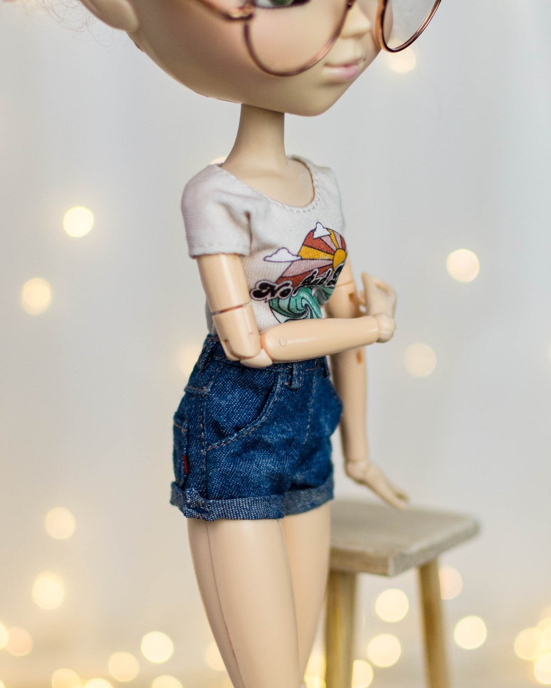 May include: A doll wearing a white t-shirt with a colorful graphic of a sun, mountains, and waves. The doll is also wearing blue denim shorts. The doll is posed with arms crossed, and is wearing glasses.