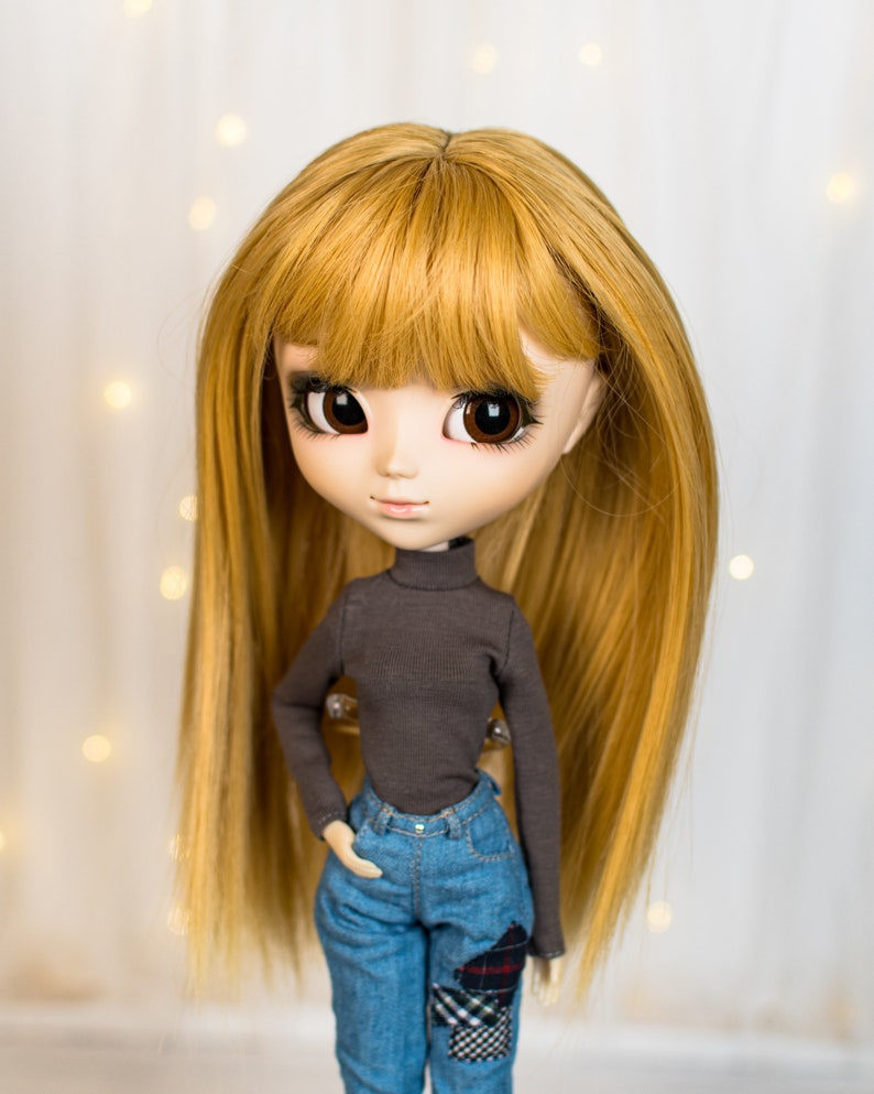 May include: A doll with long, straight, golden blonde hair, wearing a dark grey turtleneck sweater and blue jeans with a plaid patch.
