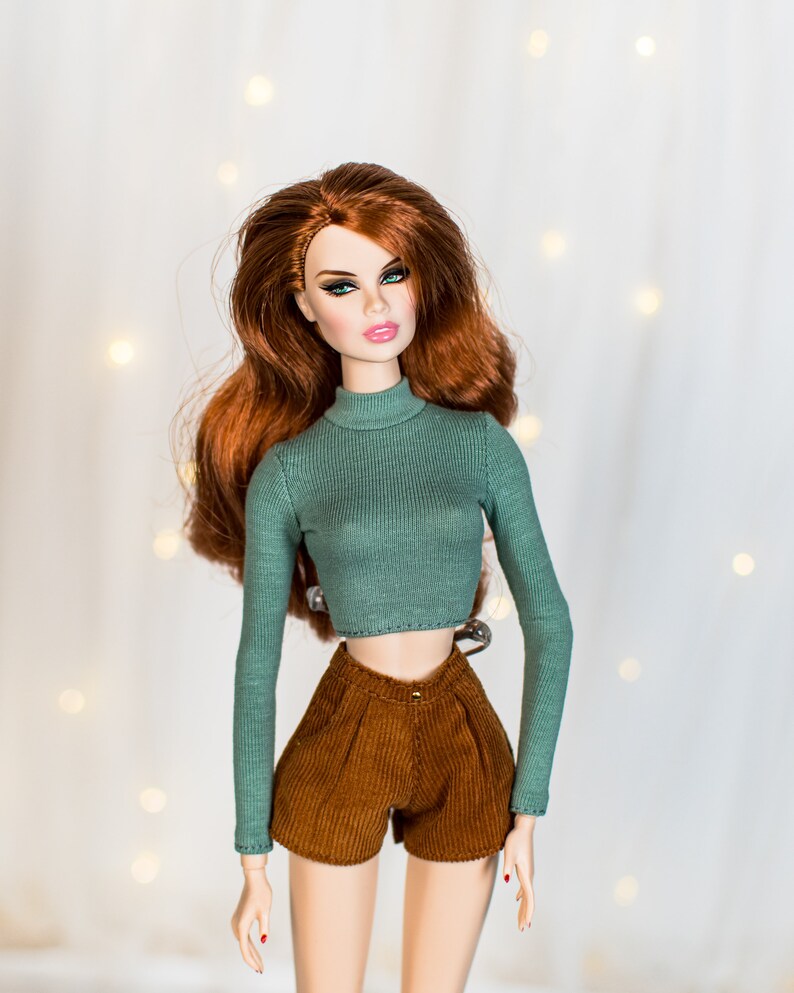 May include: A doll with long, wavy red hair wearing a green turtleneck sweater and brown corduroy shorts.