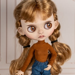May include: A doll with long, wavy, light brown hair styled in two braids. The doll is wearing a brown turtleneck sweater and blue denim shorts. The doll has brown eyes and freckles.