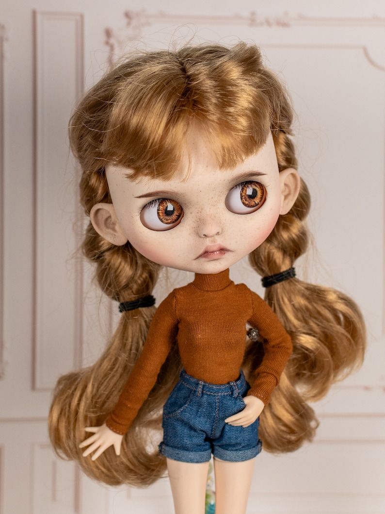 May include: A doll with long, wavy, light brown hair styled in two braids. The doll is wearing a brown turtleneck sweater and blue denim shorts. The doll has brown eyes and freckles.