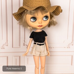 May include: A doll with blonde hair wearing a black t-shirt, beige shorts, a brown straw hat, and a black belt. The doll is standing in front of a white wall with decorative molding.