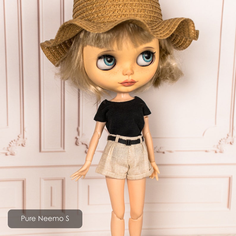 May include: A doll with blonde hair wearing a black t-shirt, beige shorts, a brown straw hat, and a black belt. The doll is standing in front of a white wall with decorative molding.