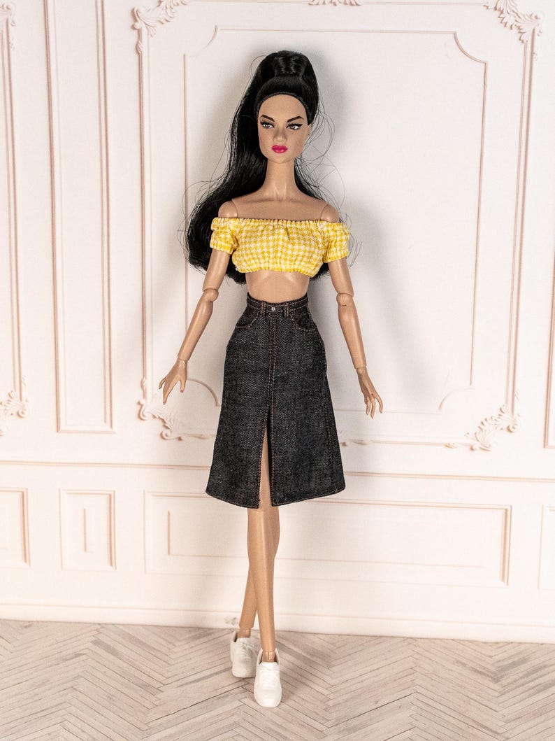 May include: A fashion doll wearing a yellow gingham crop top and a dark denim skirt with a front slit. The doll is also wearing white sneakers. The doll has long black hair and is posed in front of a white wall.