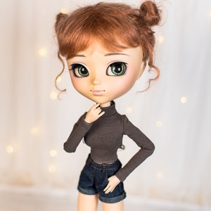 May include: A doll with red hair styled in two buns, wearing a brown turtleneck shirt and blue denim shorts. The doll has green eyes and a light skin tone.