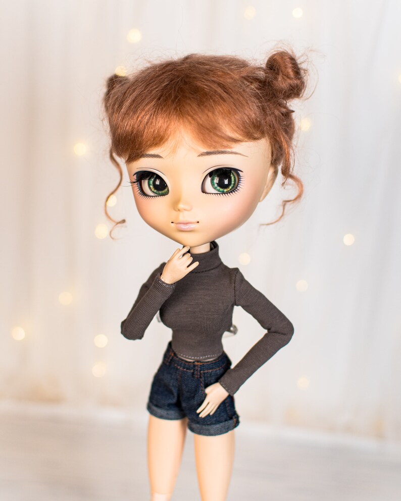 May include: A doll with red hair styled in two buns, wearing a brown turtleneck shirt and blue denim shorts. The doll has green eyes and a light skin tone.