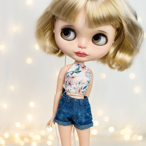 May include: A doll with blonde hair and large, expressive eyes. The doll wears a floral print halter top and denim shorts. The doll's skin has freckles, and the background is softly lit with bokeh lights.