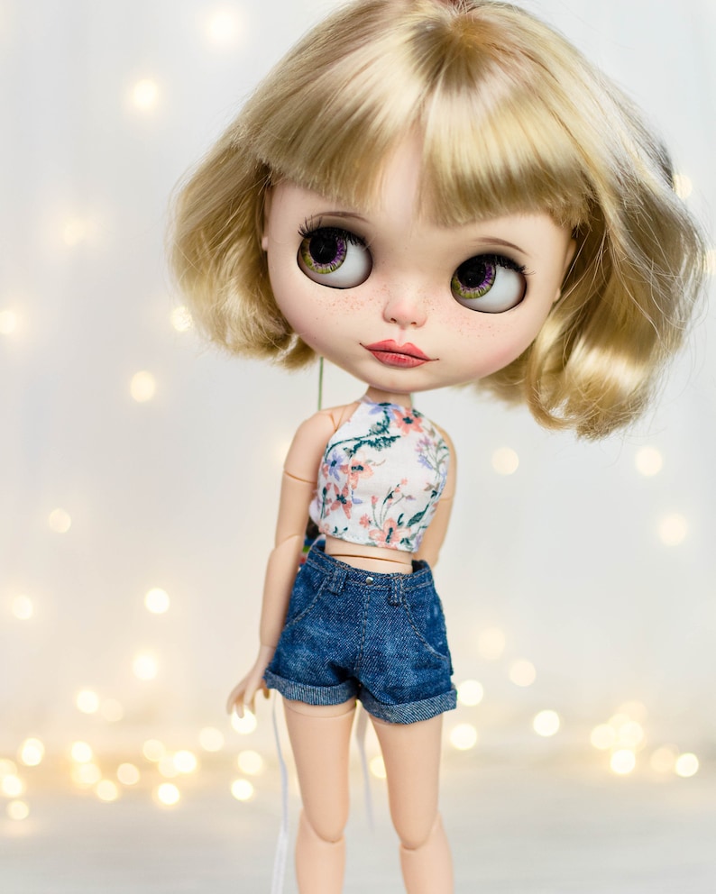 May include: A doll with blonde hair and large, expressive eyes. The doll wears a floral print halter top and denim shorts. The doll's skin has freckles, and the background is softly lit with bokeh lights.