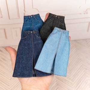 May include: A hand holds four miniature denim skirts in shades of blue and grey. Each skirt features a front slit and classic jean details, such as pockets and button closure. These skirts are designed for dolls or small figures.
