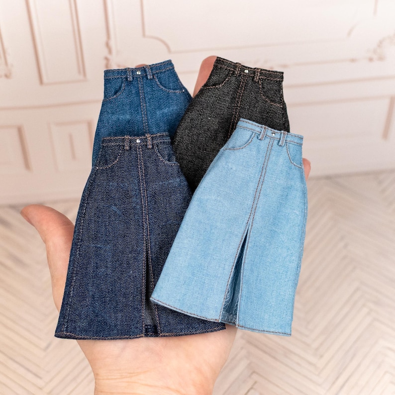 May include: Four miniature denim skirts in various shades of blue and gray are held in a hand. Each skirt has a front slit and classic jean details like pockets and a button closure. The skirts are designed for dolls or other small figures.