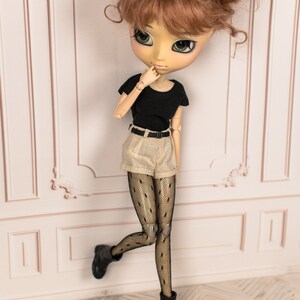 May include: A doll with brown hair styled in two buns, wearing a black t-shirt, beige shorts, black fishnet tights, and black boots. The doll is standing with one leg raised and is looking to the side.