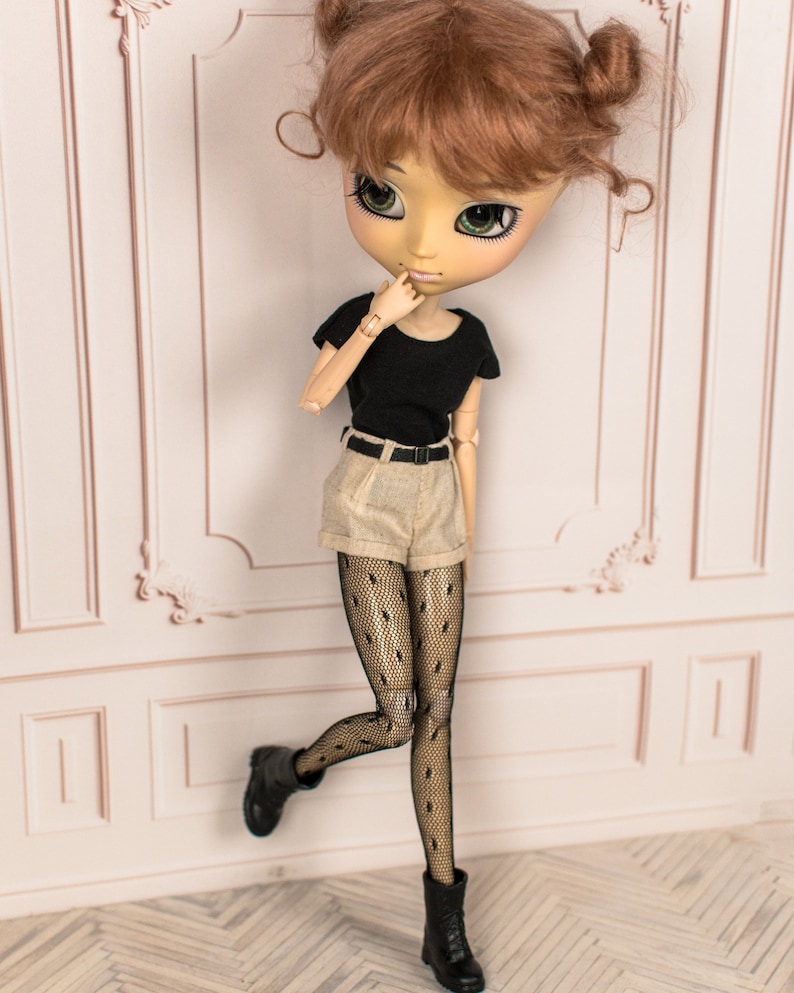 May include: A doll with brown hair styled in two buns, wearing a black t-shirt, beige shorts, black fishnet tights, and black boots. The doll is standing with one leg raised and is looking to the side.