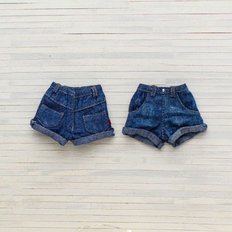 May include: Two pairs of miniature blue denim shorts with rolled-up cuffs. The shorts have pockets and belt loops, and one pair has a small red tag. The shorts are displayed on a white wooden surface.