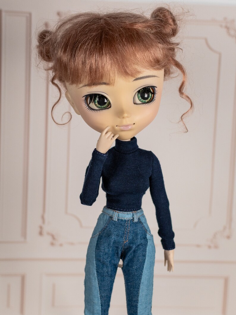 May include: A doll with brown hair styled in two buns, wearing a dark blue turtleneck sweater and blue jeans.