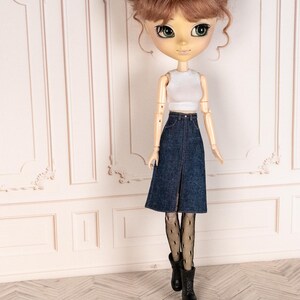 May include: A doll with brown hair styled in two buns, wearing a white crop top, a denim skirt, black fishnet stockings, and black boots. The doll has large green eyes and is posed in front of a white wall.