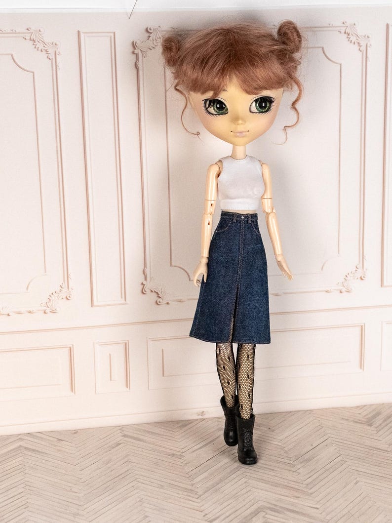 May include: A doll with brown hair styled in two buns, wearing a white crop top, a denim skirt, black fishnet stockings, and black boots. The doll has large green eyes and is posed in front of a white wall.