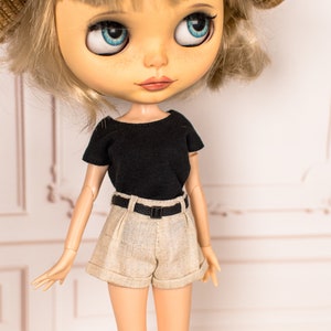 May include: A doll with blonde hair wearing a black short-sleeved shirt, beige shorts with a black belt, and a straw hat.