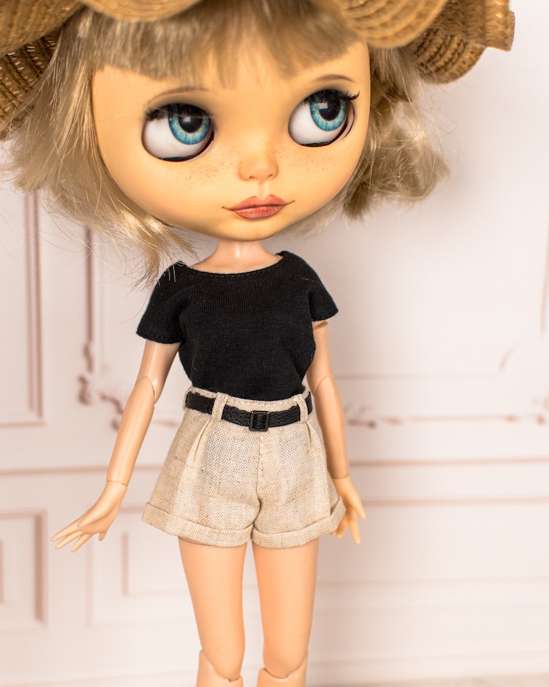 May include: A doll with blonde hair wearing a black short-sleeved shirt, beige shorts with a black belt, and a straw hat.