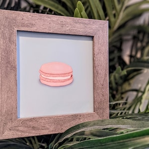 May include: A framed print of a pink macaron with white filling, set against a light blue backdrop. The artwork is presented in a square, grey wooden frame. The image is set against a backdrop of green foliage.