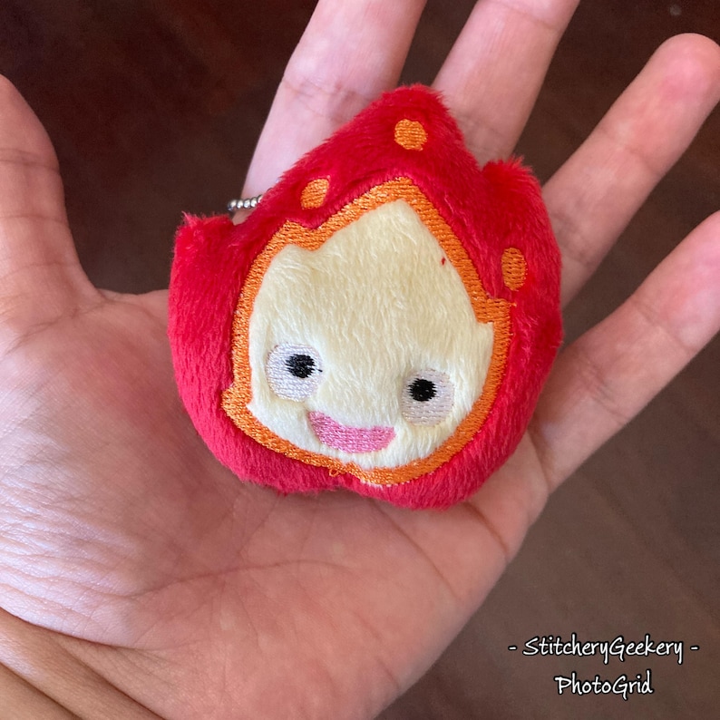 May include: A plush keychain in the shape of a cartoon fire character. The character has a red exterior with orange accents and a cream-colored face. It features embroidered black eyes and a pink smile. The keychain is attached to a silver chain.