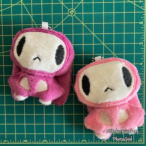 May include: Two plush keychains in shades of pink and magenta. Each keychain features a white face with black eyes and a small embroidered mouth. The plushies have long ears and a small loop for attaching to keys or a bag. The background is a green cutting mat.