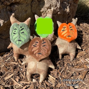 May include: Four plush figures with leaf-shaped faces in green, orange, and brown. The figures have tan bodies and arms, with embroidered facial features. The figures are arranged on a bed of wood chips, with a tree trunk in the background.