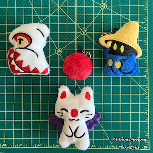 May include: A collection of plush keychains featuring characters from a video game. The keychains include a white creature with red accents, a red pom-pom, a blue mage with a yellow hat, and a white cat with purple wings. The keychains are on a green cutting mat.