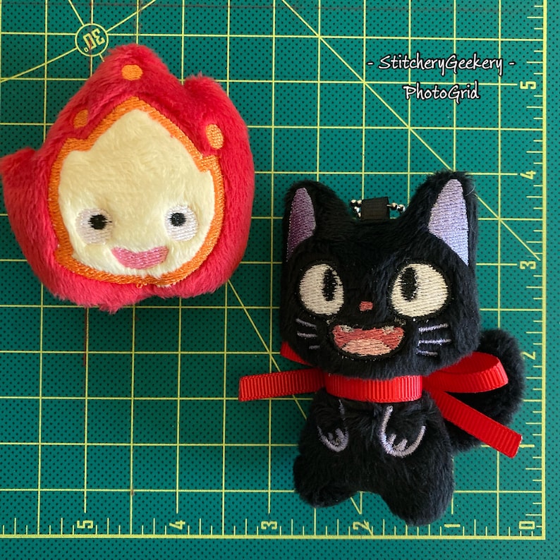 May include: Two plush keychains: a red and yellow fire spirit and a black cat with a red bow. The fire spirit has a smiling face. The cat has purple ears and a smiling face. The keychains are on a green grid.