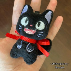 May include: A plush black cat keychain with embroidered details. The cat has large, round eyes, a smiling mouth, and a red ribbon tied around its neck. The cat's ears are purple. The keychain is held in a hand.