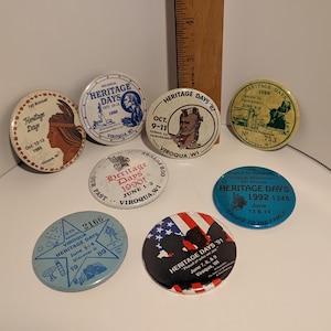 May include: A collection of nine round, colorful buttons with the text "Heritage Days" and various years, dates, and locations. The buttons are a mix of blue, red, white, yellow, and green.