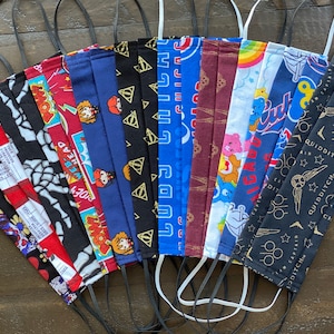 Könnte beinhalten: Eine Sammlung von 14 bunten Stoffmasken mit verschiedenen Designs, darunter Totenköpfe, Skelette, Harry Potter, Care Bears und Quidditch. Die Masken haben schwarze elastische Ohrschlaufen.