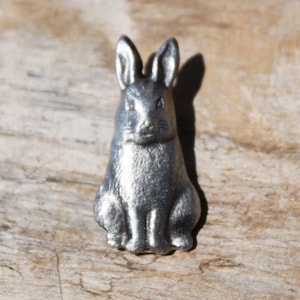 May include: A small, silver-colored rabbit figurine. The bunny is sitting upright with its ears perked up. The figurine is made of metal and has a detailed texture. The background is a light brown, wooden surface.