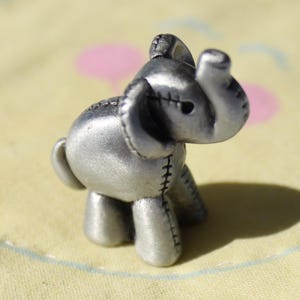 May include: A small, silver-colored elephant figurine with stitched details. The elephant is standing on a yellow fabric with a sun-like design and a pink heart. The figurine's shadow is visible on the fabric.