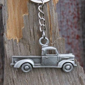 May include: A silver metal keychain featuring a vintage pickup truck. The truck has a flatbed and a detailed design. The keychain is hanging from a silver chain and ring.