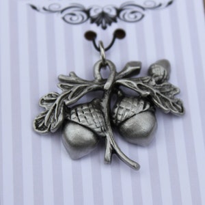 May include: A silver charm pendant featuring two acorns with leaves. The acorns are detailed with ridges and the leaves are delicate and realistic.