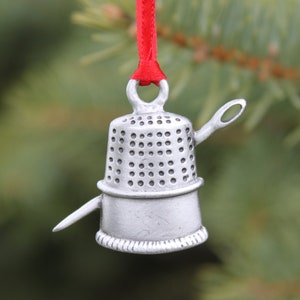 May include: A silver thimble Christmas ornament with a needle attached. The ornament is hanging from a red ribbon.