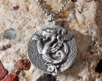 May include: Silver necklace with a round pendant featuring a detailed, three-dimensional winged dragon design.