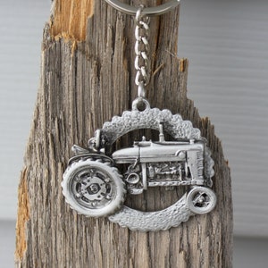 May include: A silver metal keychain featuring a detailed tractor design. The tractor is surrounded by a circular, textured border. The keychain is attached to a silver chain and a silver key ring.