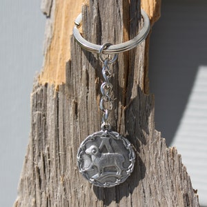 May include: Silver keychain with a circular charm featuring a ram or sheep zodiac symbol. The charm is surrounded by a decorative border of stars and thorns.