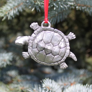 May include: A silver turtle ornament with a detailed shell design. The ornament is hanging from a red ribbon.