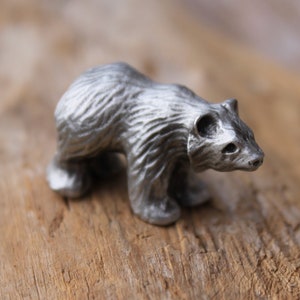 May include: A small, silver-colored bear figurine. The bear is depicted in a walking pose, with detailed fur texture. The figurine is set against a wooden background, highlighting its metallic finish. A decorative item.