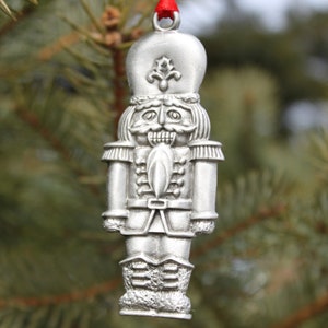 May include: A silver-colored nutcracker ornament with a red ribbon hanging from a Christmas tree. The nutcracker is wearing a hat with a fleur-de-lis design.
