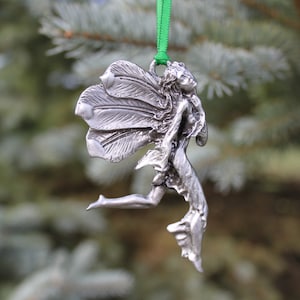 May include: A silver pewter ornament of a fairy with wings outstretched, flying with one leg extended. The ornament is hanging from a green ribbon.