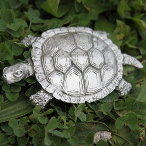 May include: A silver turtle figurine with a detailed shell design, resting on a bed of green leaves.