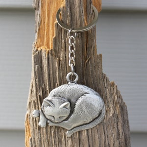 May include: A silver metal keychain featuring a sleeping cat design. The keychain is attached to a silver chain and ring.