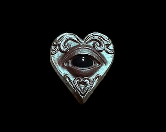 May include: A silver heart-shaped pendant with an eye design. The eye is black and the heart is decorated with intricate scrollwork.