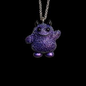 May include: A purple monster pendant with a silver chain. The pendant has a textured surface, small black horns, and a smiling face. The monster's body is a vibrant purple with hints of blue and pink.