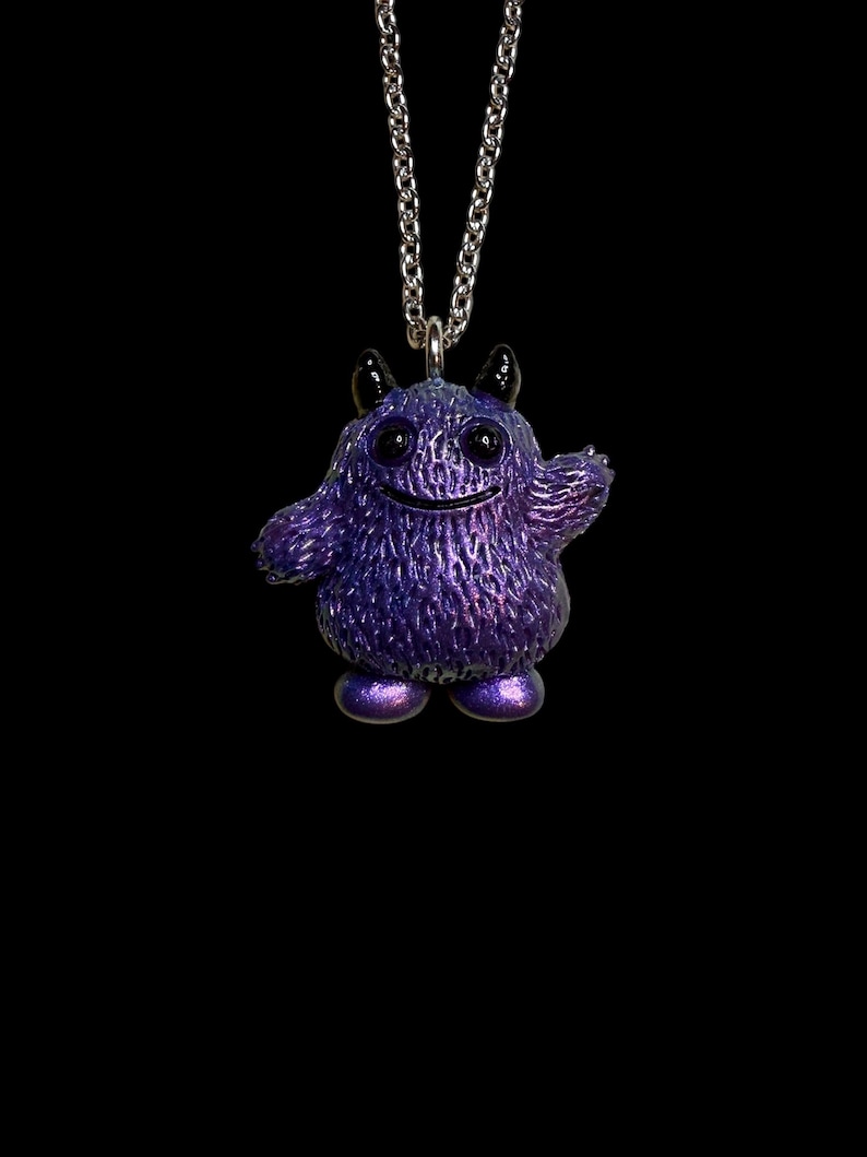 May include: A purple monster pendant with a silver chain. The pendant has a textured surface, small black horns, and a smiling face. The monster's body is a vibrant purple with hints of blue and pink.