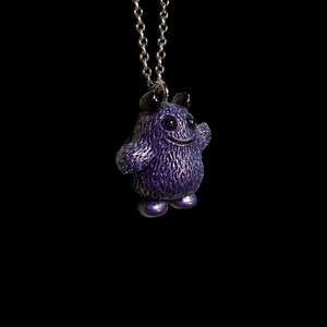 May include: A purple monster pendant on a silver chain. The pendant has a textured surface, small horns, and a smiling face. The monster is standing with its arm raised. The background is black.